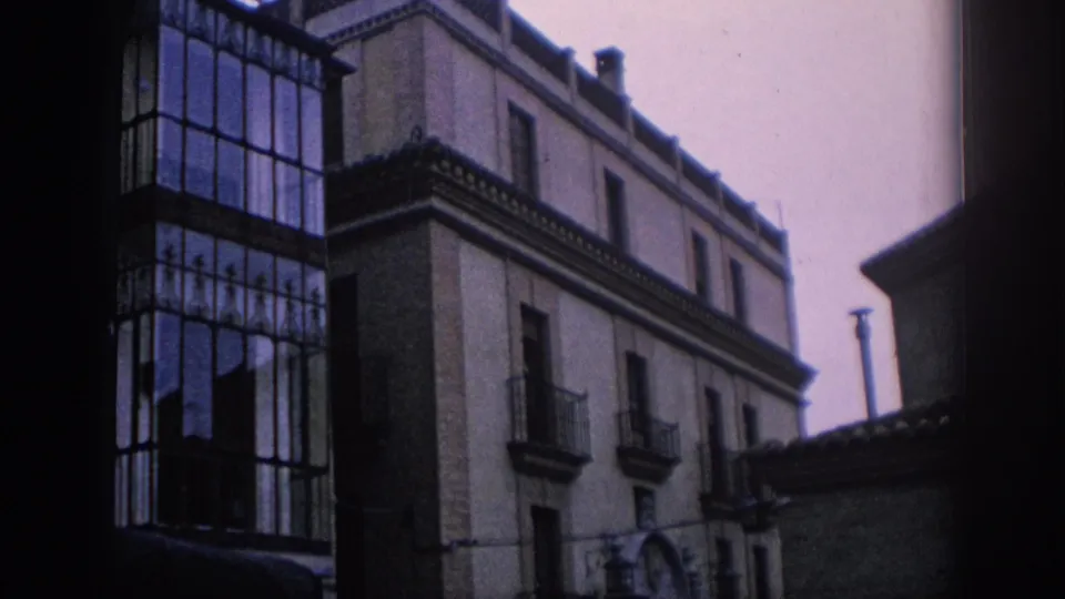 1966 Spain home-movie frame of an urban facade with a tall glass balcony enclosure, small balconies, and soft faded color.