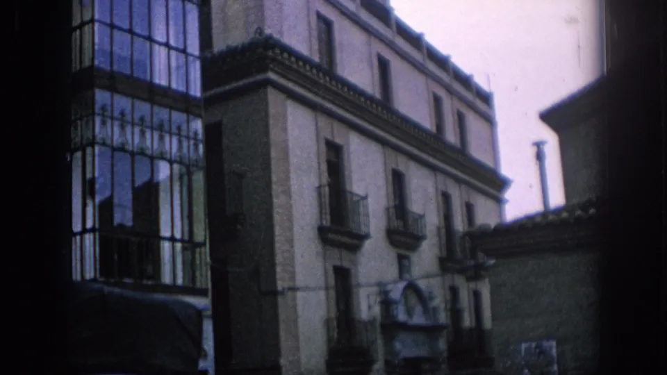 1966 Spain street view of a multi-story building with enclosed glass balcony, iron railings, and neighboring roof edges.