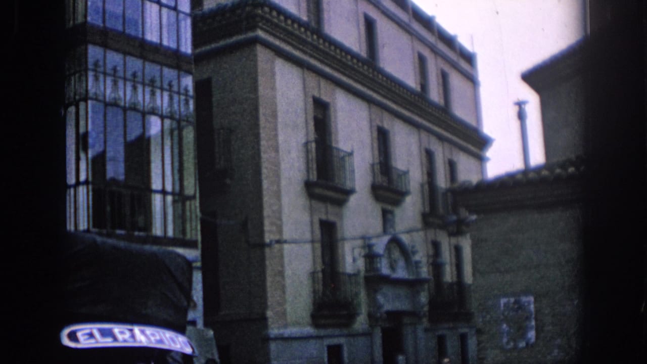 1966 Spain home-movie frame showing old town buildings, iron balconies, and the front of a vehicle with visible lettering.