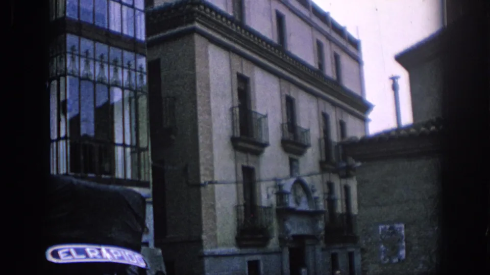 1966 Spain home-movie frame showing old town buildings, iron balconies, and the front of a vehicle with visible lettering.