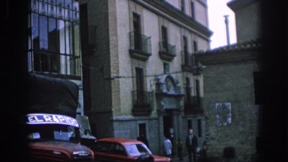 1966 Spain street frame with old facades, a red sedan, a dark vehicle with signage, and people near a doorway.