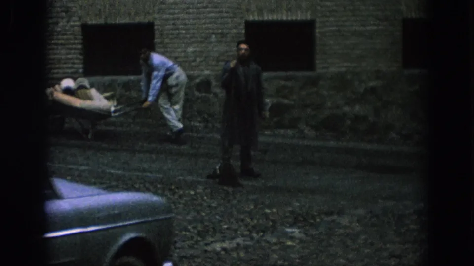1966 Spain home-movie frame showing a man in a dark coat on a stone sidewalk as a worker handles a wheelbarrow nearby.