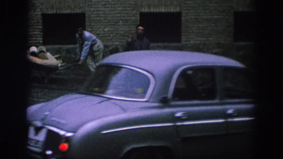 1966 Spain street frame with a gray sedan moving past a worker, a wheelbarrow, and a man standing near a weathered brick wall.