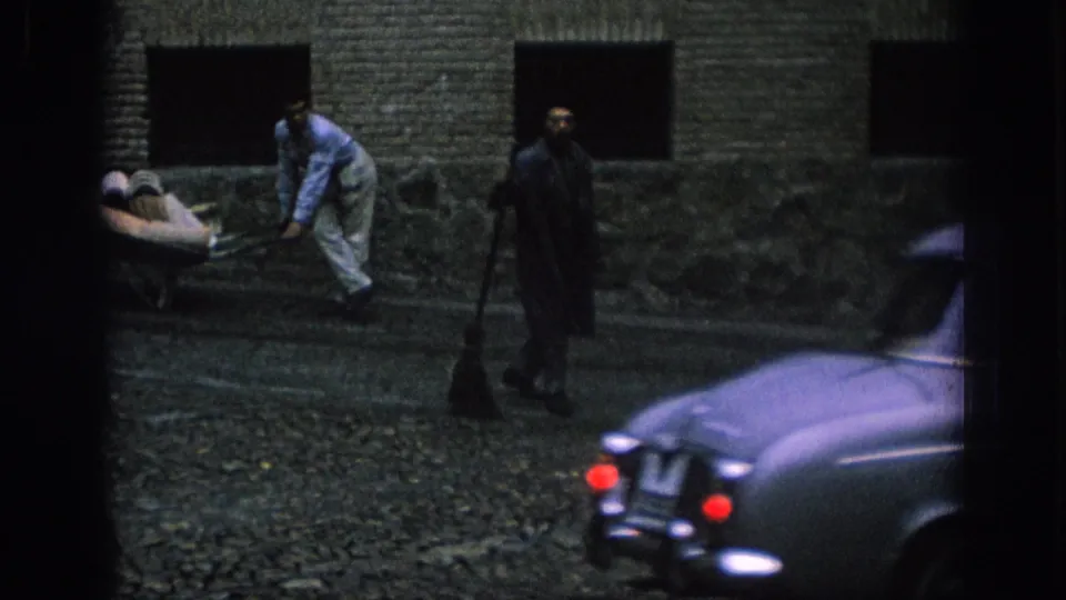 In Spain, 1966, two men appear to clean a cobblestone street beside a wall while a rounded mid-century car sits partly in frame.