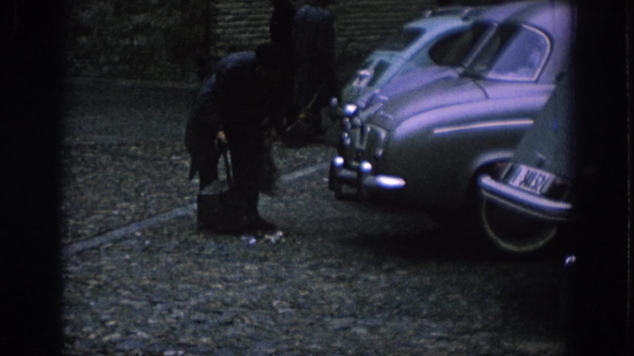 In Spain, 1966, a man bends toward a small container on a cobblestone street between parked mid-century cars.