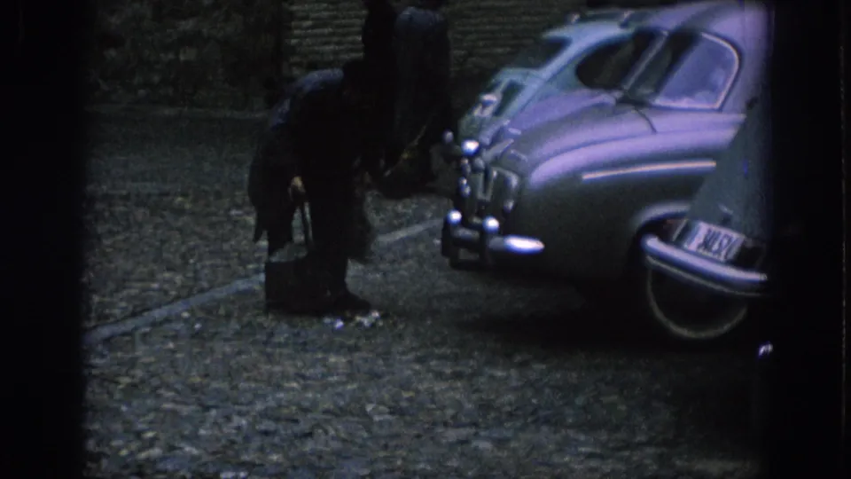 In Spain, 1966, a man bends toward a small container on a cobblestone street between parked mid-century cars.