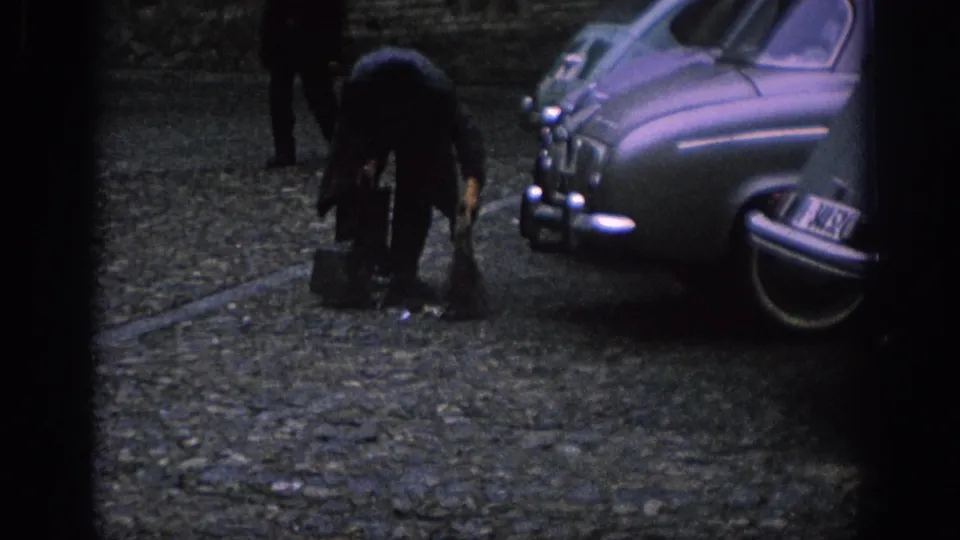 In Spain, 1966, a man leans down near a small container while classic rounded cars line the cobblestone street.
