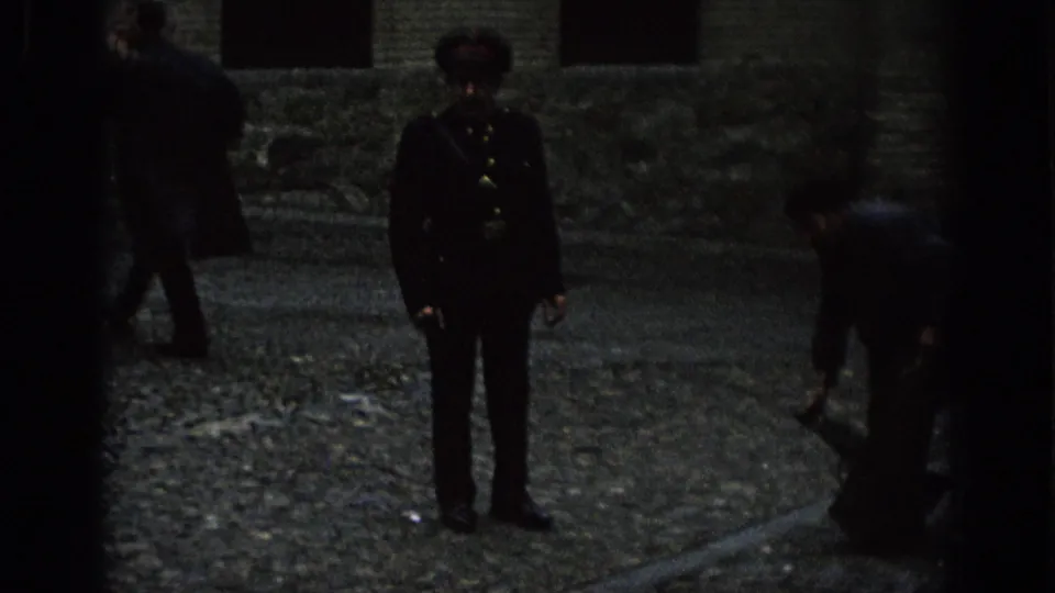 In 1966 Spain, a uniformed guard stands in a dim cobblestone area as nearby people handle luggage beside parked vehicles.