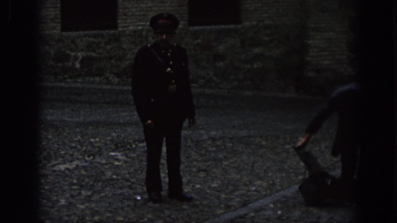 Uniformed guard stands near a person bending over luggage on a dim cobblestone surface.