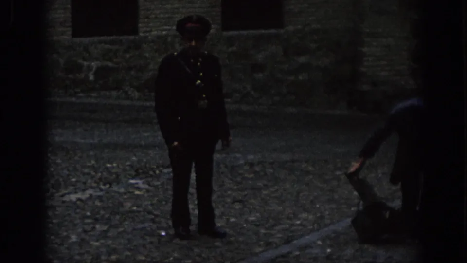A uniformed guard remains centered in a dim Spanish cobblestone setting while a person bends over luggage near the right edge.