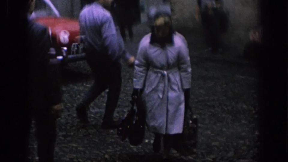 In a 1966 Spanish street or parking area, a woman in a light coat and dark head covering walks with bags near a red car.