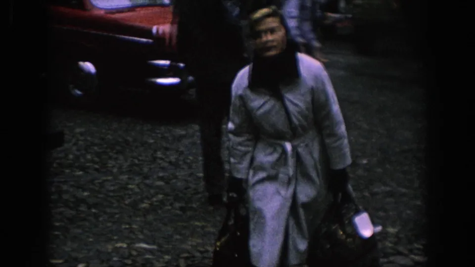 A woman in a light coat and dark headscarf walks toward the camera with bags near a red car in 1966 Spain.