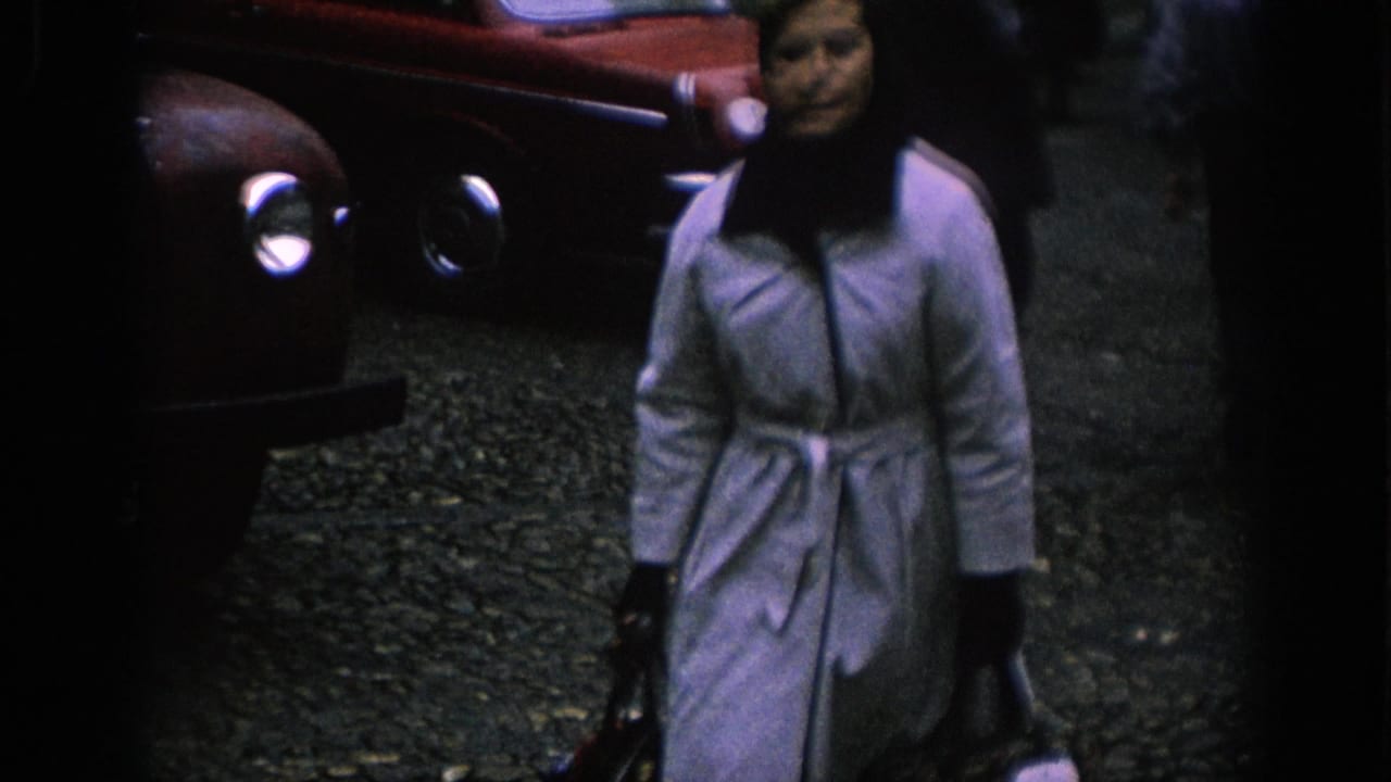 In 1966 Spain home-movie footage, a woman carrying bags walks past red classic cars on a cobblestone street.