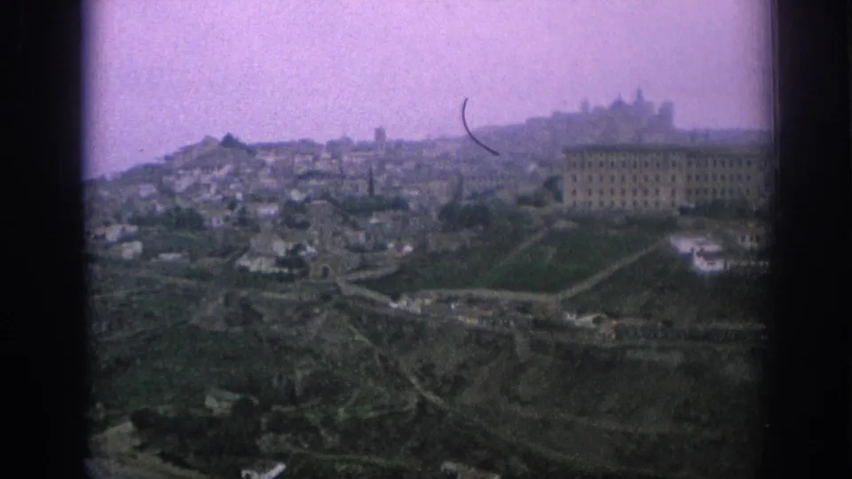 1966 Spain 8mm frame showing a hazy elevated panorama of a hillside city with terraced slopes and a prominent institutional-looking building.