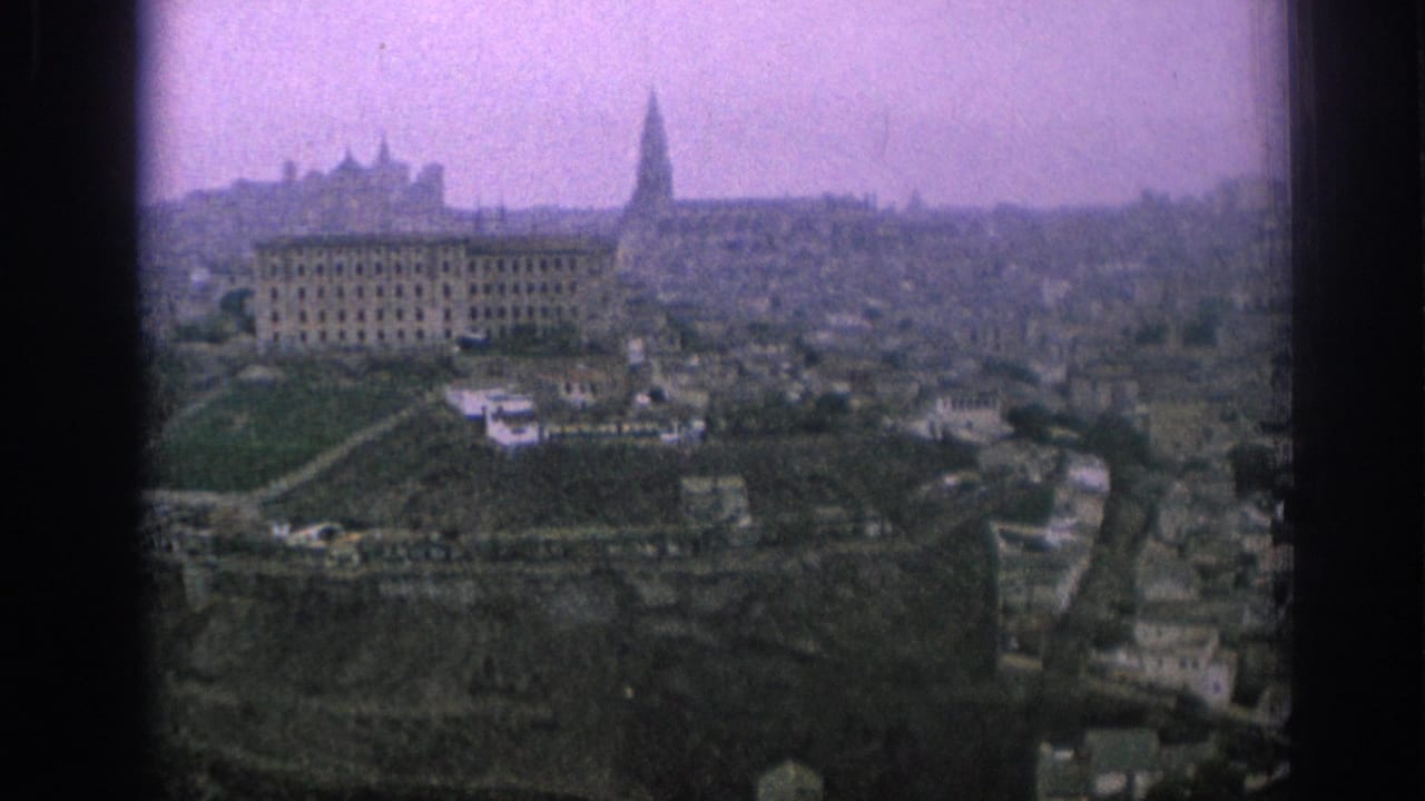 1966 Spain travel-film frame with a broad city view, terraced hillside paths, pale buildings, and a church-like spire in the distance.