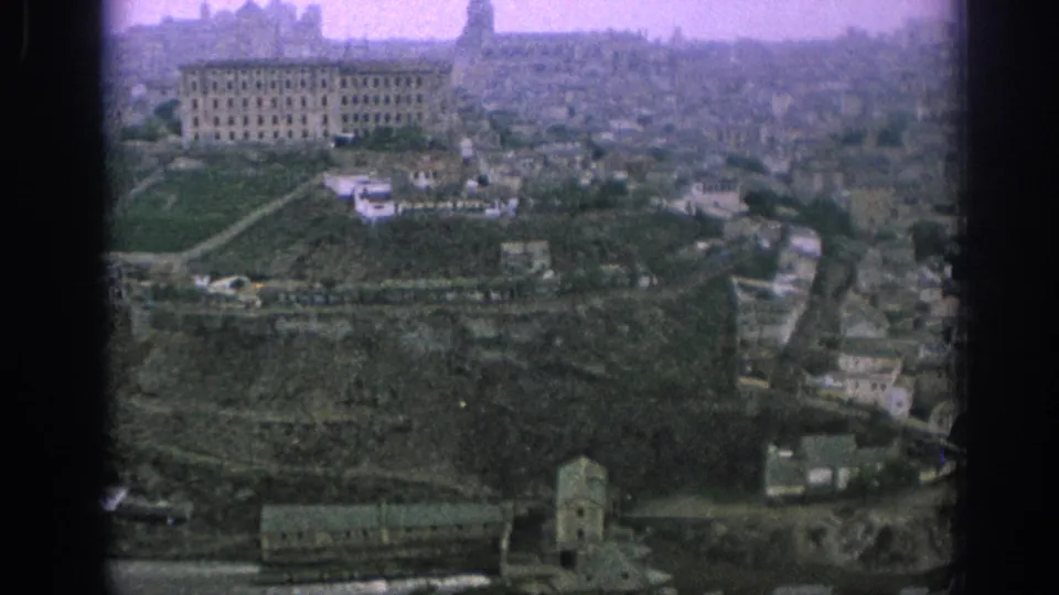 1966 Spain 8mm frame looking across a sloped urban edge with terraces, pale buildings, roads, trees, and a waterway below.