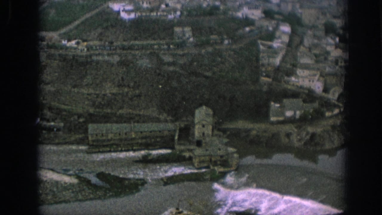 1966 Spain 8mm frame showing an elevated view over a historic riverside settlement, watercourse, and large stone structure near the bank.
