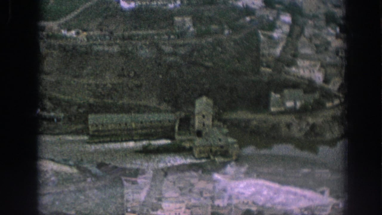 1966 Spain 8mm frame with a shifted elevated view of riverside stone buildings, water, and hillside development.