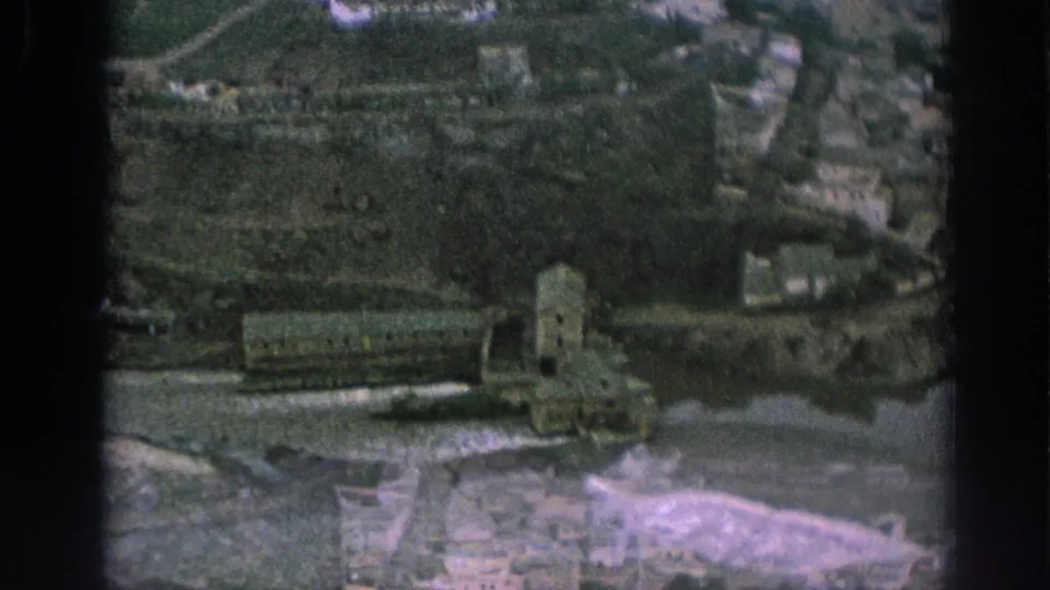 1966 Spain 8mm frame with a shifted elevated view of riverside stone buildings, water, and hillside development.