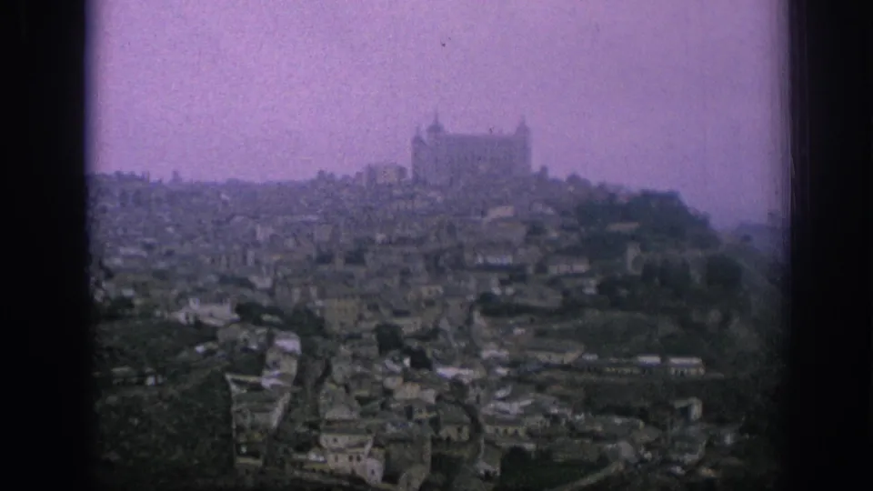 1966 Spain 8mm frame showing a hazy hillside town below a large cathedral or castle-like landmark on higher ground.