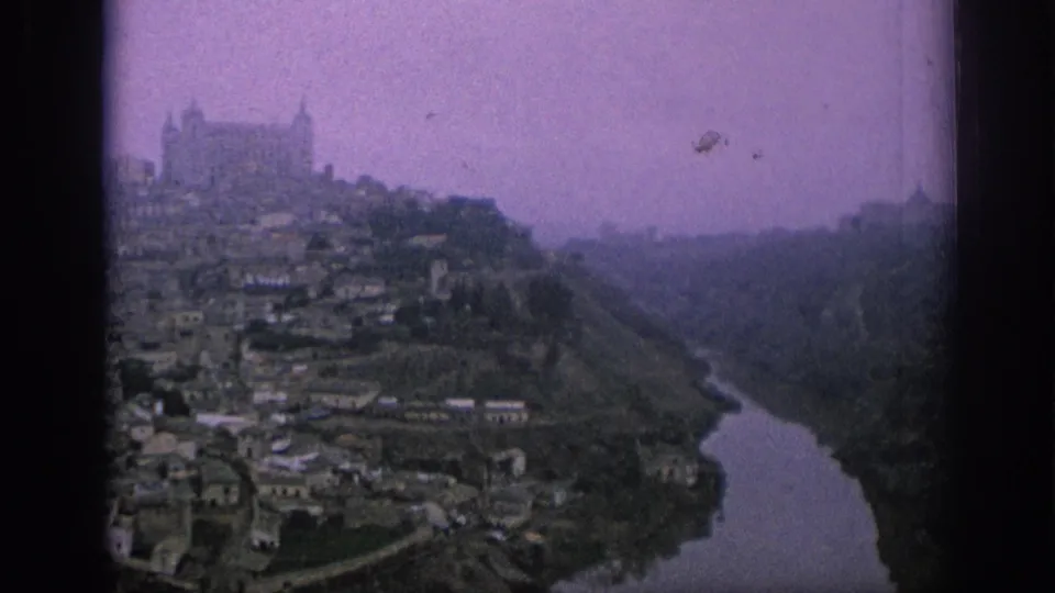 1966 Spain home-movie frame with a purple cast, showing the church skyline above a riverside hillside town.