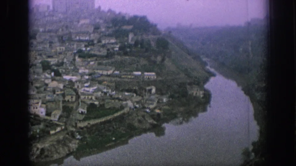 1966 Spain 8mm frame showing clustered hillside houses, a river bend, terraced slopes, and a castle-like form near the top of town.