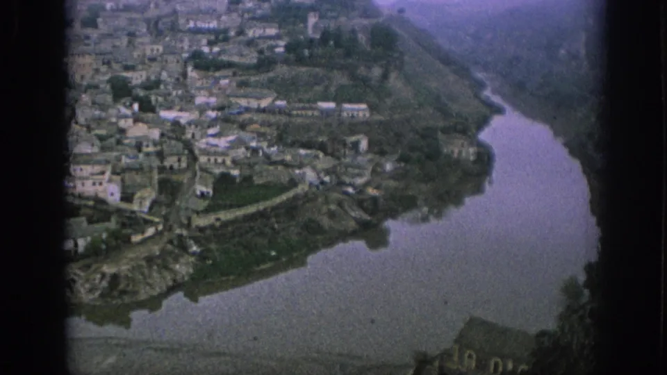 1966 Spain 8mm view of pale hillside buildings and vegetation above a broad river curve, with soft focus and aged color cast.