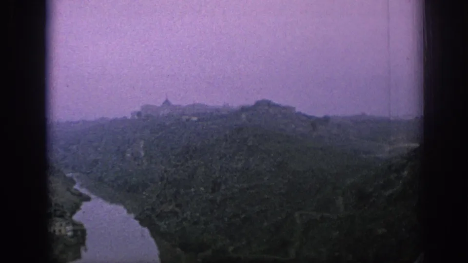 Wide 1966 Spain 8mm frame of a hilltop town and winding river valley, with the settlement receding into haze and visible film-frame edges.