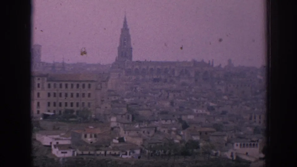 1966 Spain home-movie frame showing a tall spire rising over a dense historic town, with soft focus, dust marks, and purple tint.