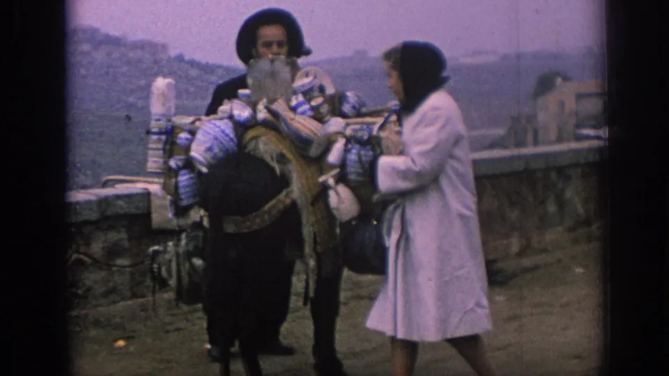 1966 Spain travel footage shows a woman near a stone railing speaking with a local seller beside a pack animal carrying bundled crafts.