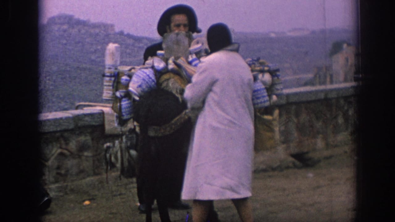 In 1966 Spain, a woman in a white coat and headscarf faces a bearded man beside a donkey carrying bundles and containers near a stone wall.