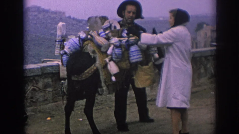 In 1966 Spain, the bearded man and woman stand close to a heavily loaded donkey, with containers and bundles hanging from the animal's pack.