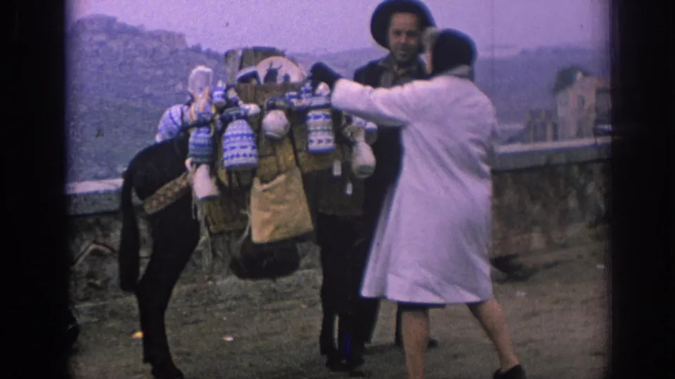 In 1966 Spain, the woman reaches toward the packed items on the donkey while the bearded handler stands beyond the animal near a stone wall.