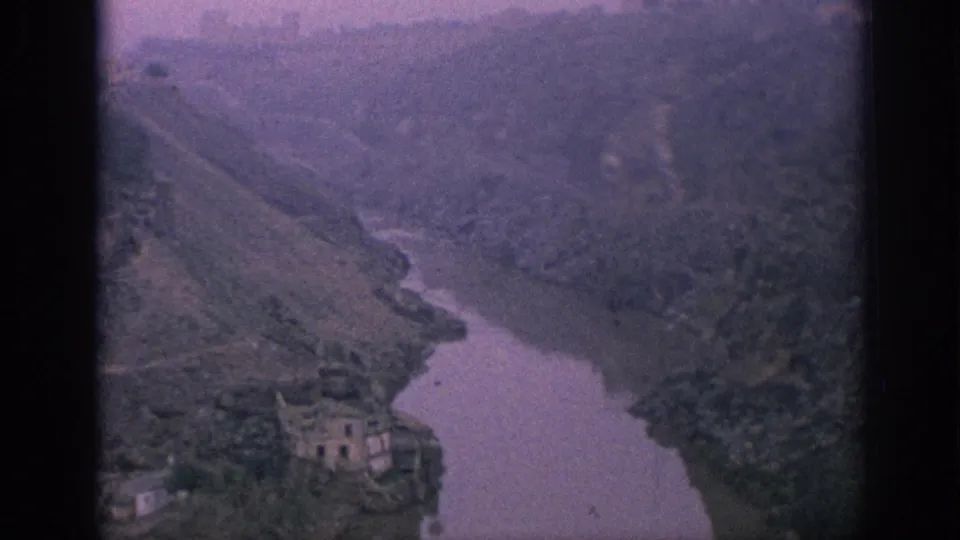 1966 Spain 8mm frame looking down on a river valley, dense hillside vegetation, and a small building near the water.