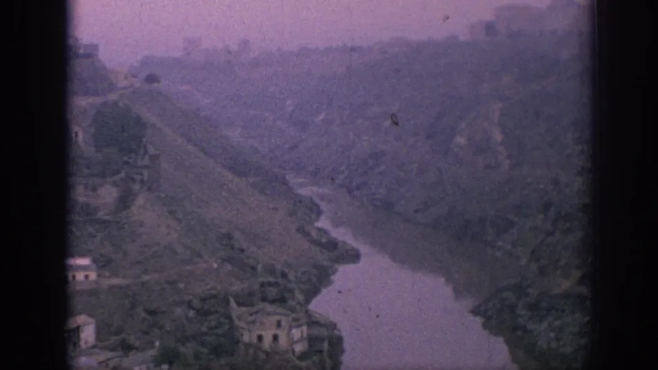 1966 Spain 8mm frame with a purple-tinted river valley, steep hills, scattered buildings, and soft distant haze.
