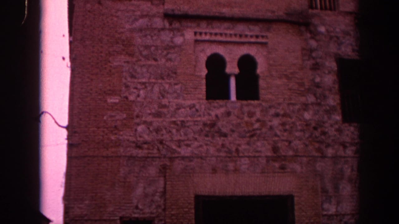 1966 Spain home-movie frame showing a historic stone and brick facade with paired arched windows and heavy magenta film tint.