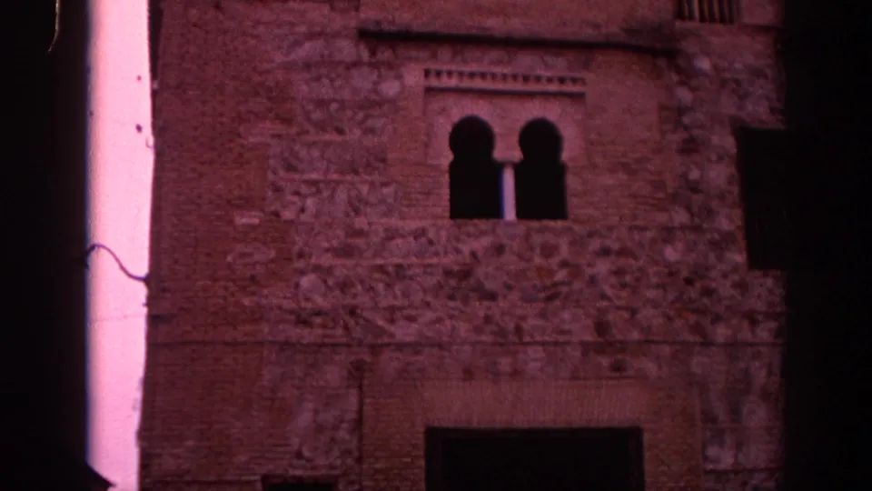 1966 Spain home-movie frame showing a historic stone and brick facade with paired arched windows and heavy magenta film tint.