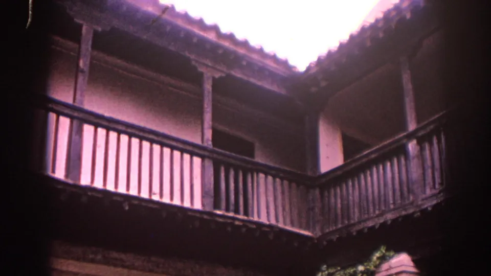 1966 Spain home-movie frame looking upward inside a courtyard toward a wooden balcony, vertical railings, and bright roofline.