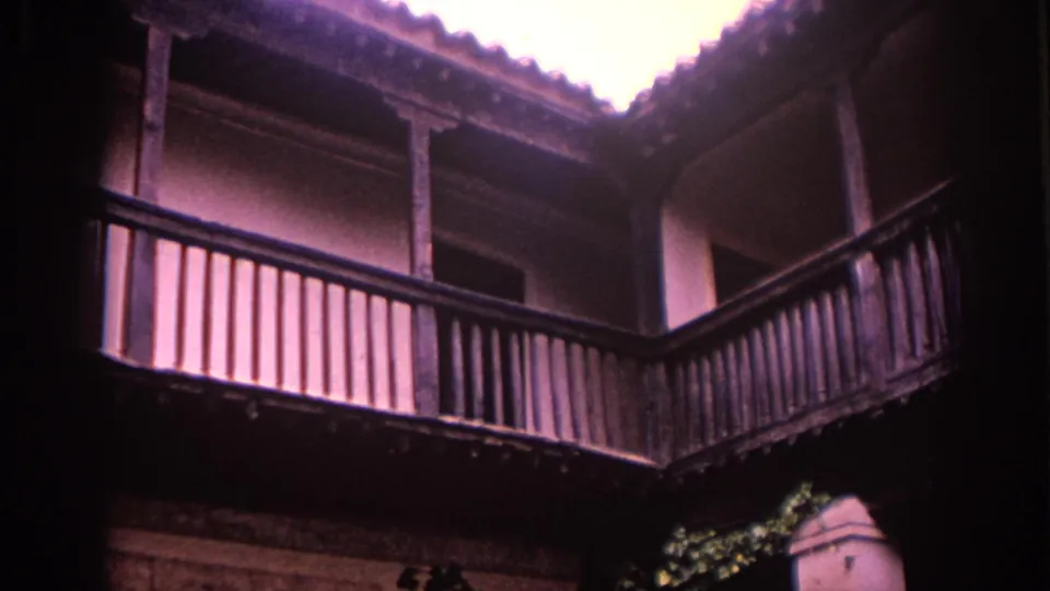 8mm frame from Spain showing a traditional courtyard veranda with dark wooden railings, stucco walls, and roof tiles.