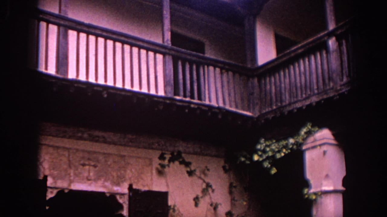 1966 Spain home-movie frame with wooden balcony rails above a plaster courtyard wall, ivy, and a dark doorway below.