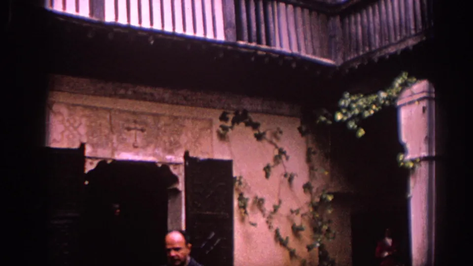 1966 Spain frame showing a courtyard doorway, open shutters, ivy, tile trim, and a partially visible person near the lower edge.