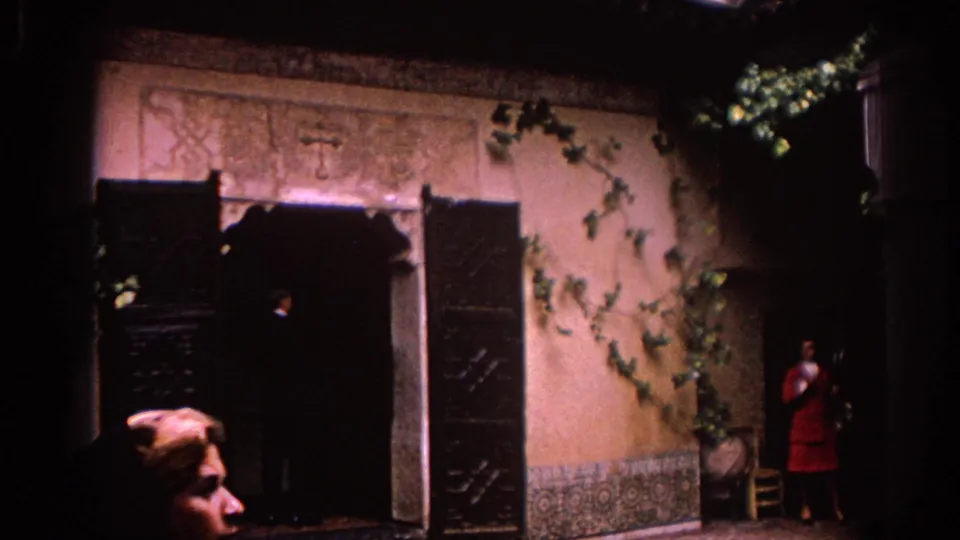 1966 Spain 8mm frame of a courtyard doorway with open shutters, ivy on the wall, and a woman in red standing near the right edge.