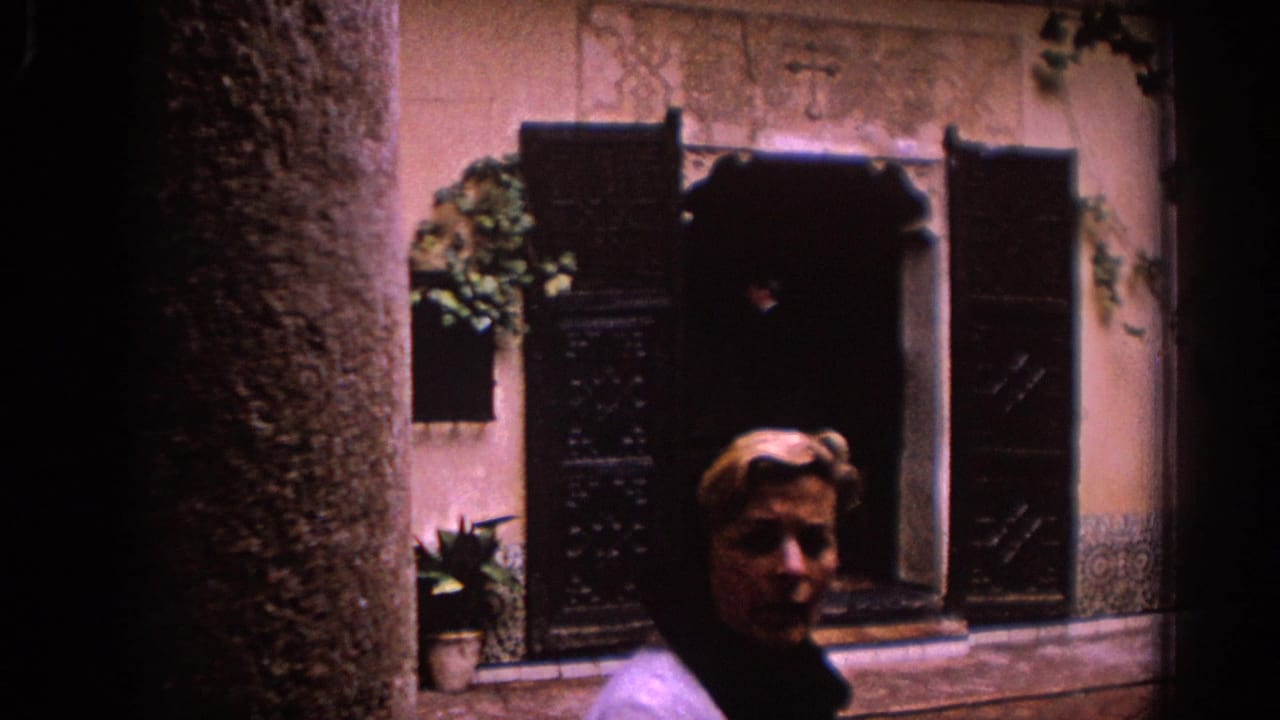 A woman in a dark top stands near the camera as shuttered windows, stucco, plants, and a shadowed courtyard wall fill the background.