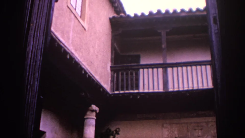 The handheld camera tilts upward to a wooden balcony, roof tiles, stucco walls, and a stone column inside the courtyard.
