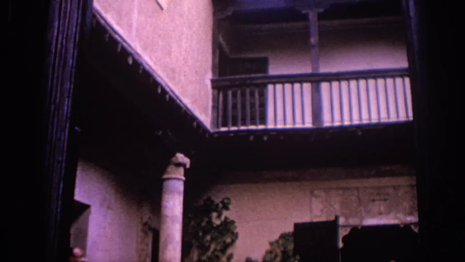 A Spanish courtyard view shows an upper wooden balcony, stone column, stucco facade, dark shutters, and greenery below.