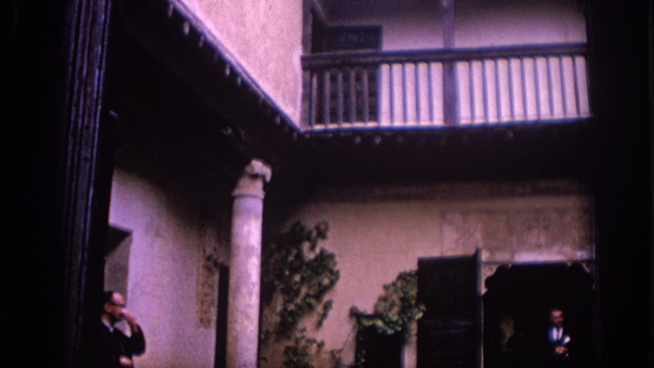 1966 Spain 8mm frame showing a traditional courtyard with a stone column, tiled wall base, climbing plants, and suited men by doorways.