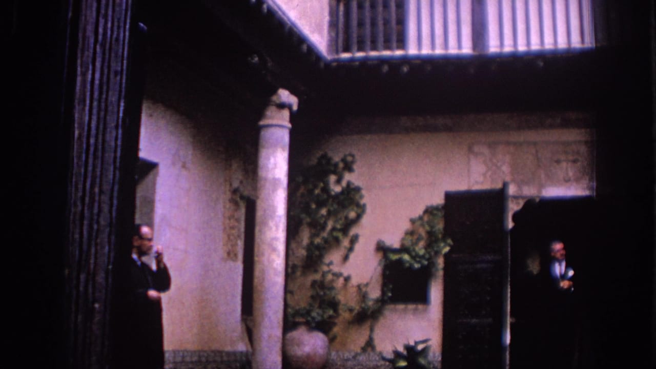 1966 Spain courtyard frame with balcony railings above, a pale stone column, plants, and suited men standing in separate doorways.