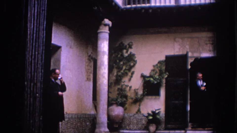 1966 Spain 8mm frame of a man smoking near a stone column, with another suited man standing in a dark doorway across the courtyard.