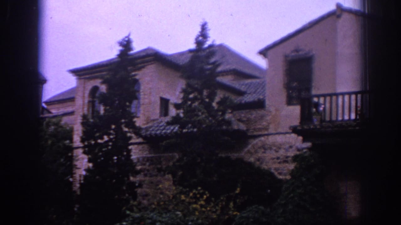 Stone and stucco building with tiled roofs, balcony, trees, and dark frame edges.