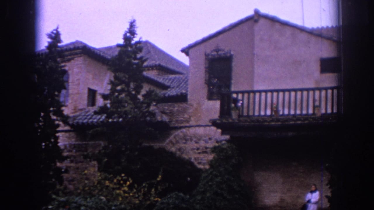 1966 Spain home-movie frame with balcony, stone wall, garden plants, dark window-like edges, and a woman partly visible at lower right.